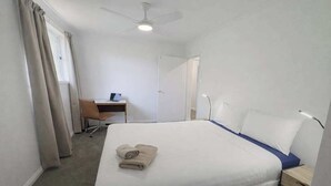 Ett hotellrum med en säng, ett skrivbord, en stol, en takfläkt och ett fönster med gardiner.