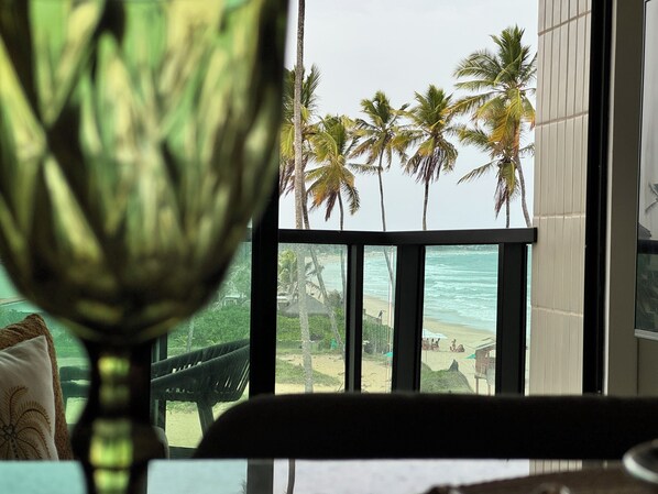 Blick aus einem Fenster auf einen Strand mit Palmen, einem Stuhl und einem Tisch.