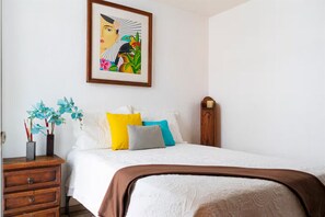 A bedroom with a bed, pillows, a nightstand, a framed artwork, and a wooden nightstand.