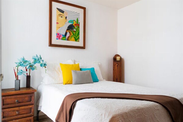 Un dormitorio con una cama, almohadas, una mesita de noche, una obra de arte enmarcada y otra mesita de noche de madera.