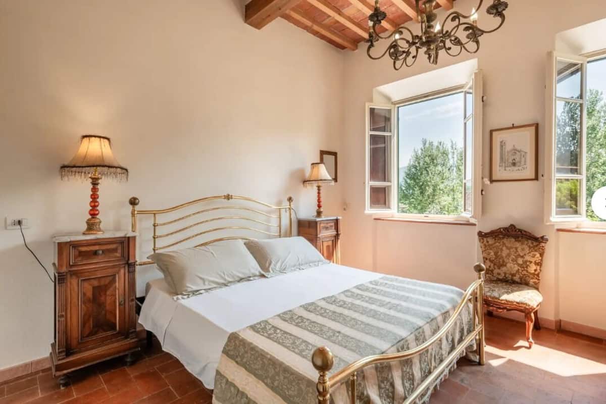 Un dormitorio con cama, lámparas de noche, un armario de madera, una silla y una ventana con vistas.