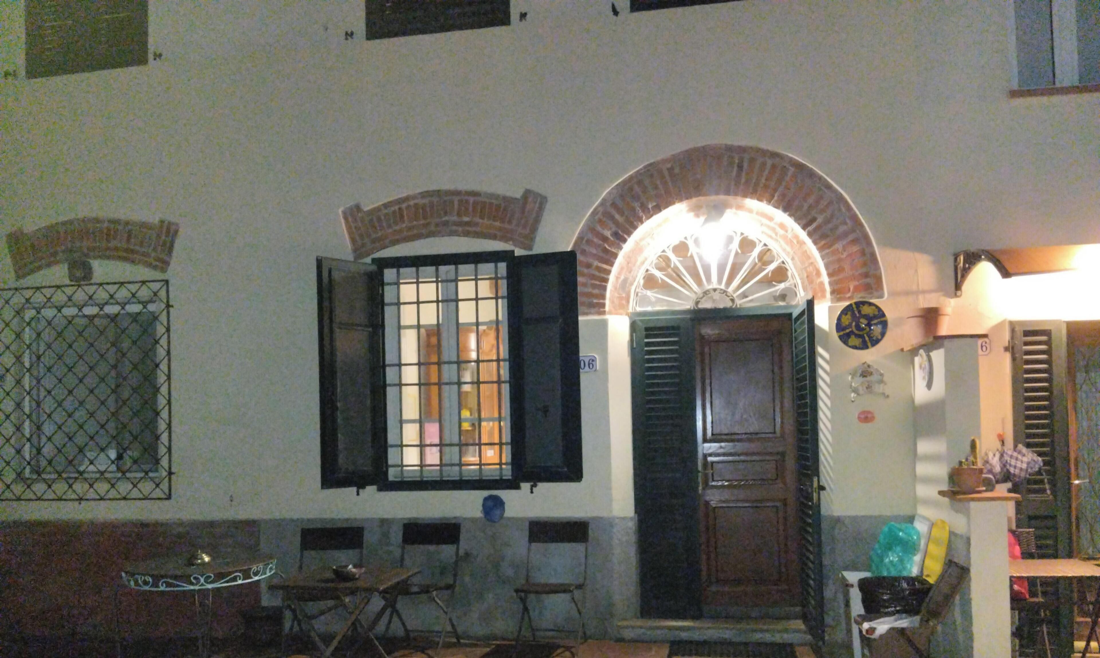 Una habitación con puerta de madera, una ventana con contraventanas abiertas, una mesa pequeña con sillas y un reloj de pared.