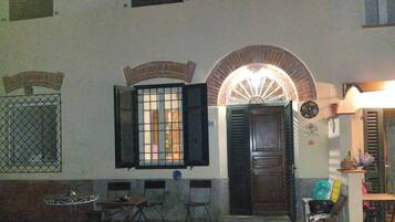 Una habitación con puerta de madera, una ventana con contraventanas abiertas, una mesa pequeña con sillas y un reloj de pared.