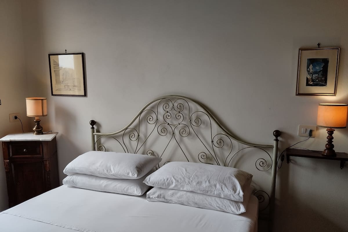 Una cama bien hecha con sábanas y almohadas blancas, un cabecero de metal con adornos en volutas, una mesita de noche con lámpara y un marco de cuadro colgado en la pared.