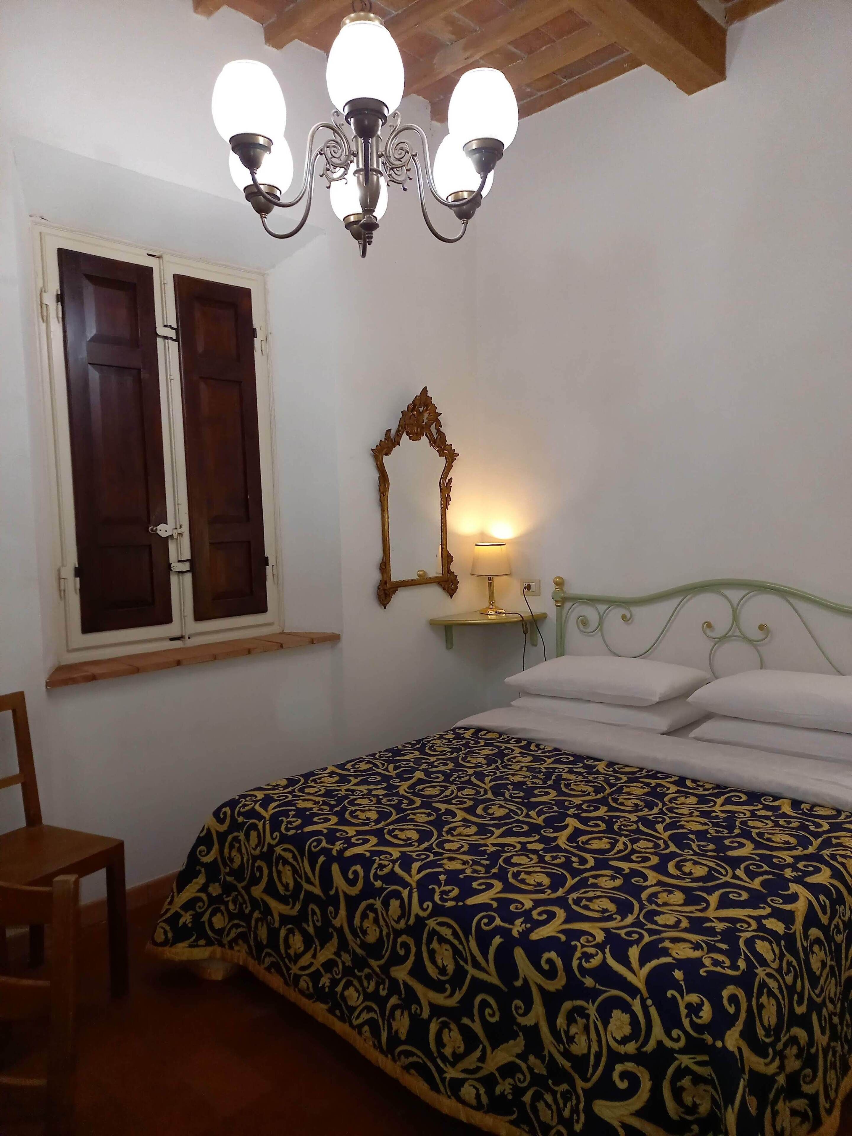 A bedroom with a bed, a wooden chair, a window with shutters, a wall-mounted mirror, and a chandelier.