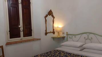 A bedroom with a bed, a wooden chair, a window with shutters, a wall-mounted mirror, and a chandelier.
