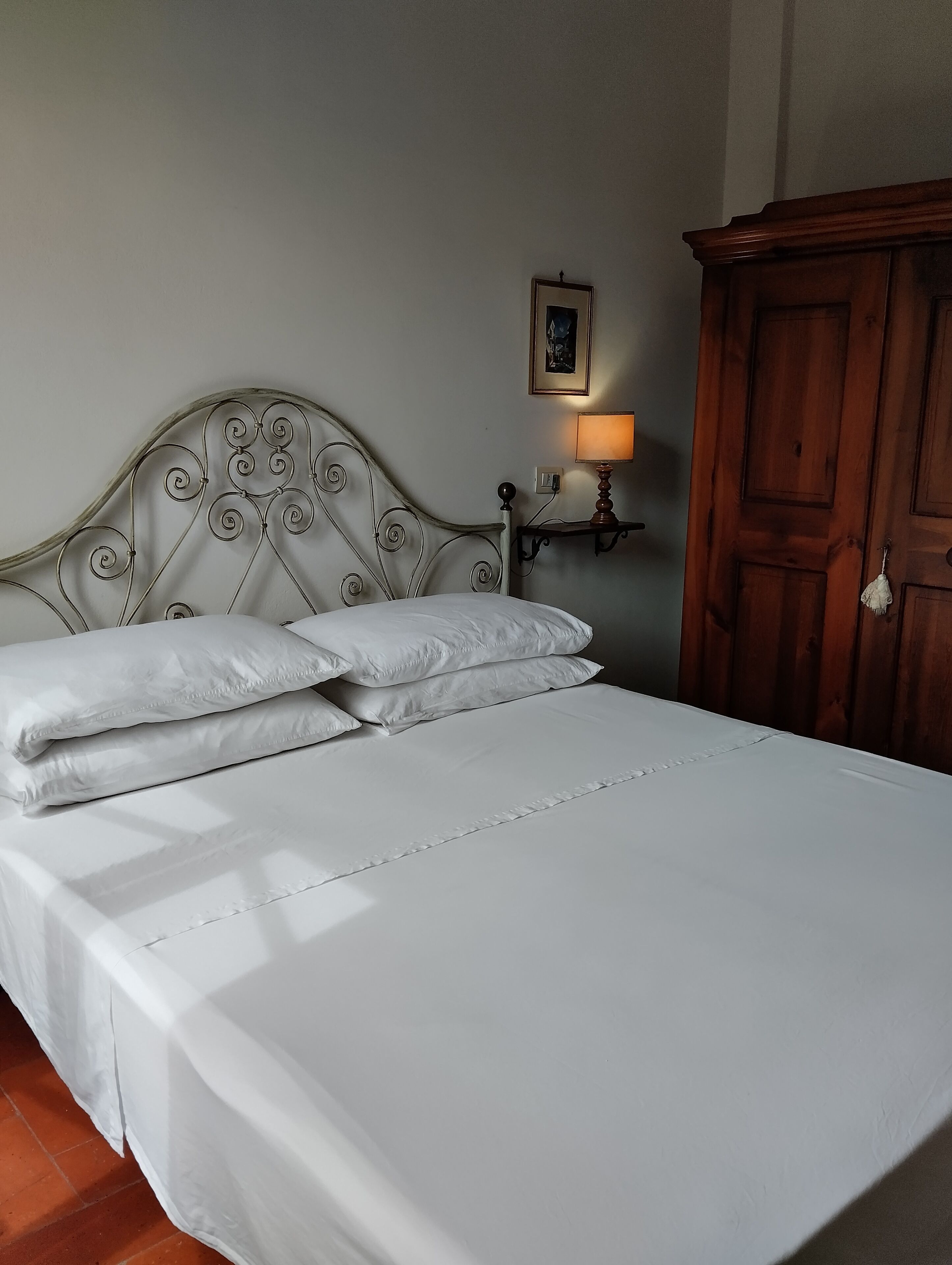 A neatly made bed with white linens, a metal headboard, a bedside table with a lamp, and a wooden wardrobe.