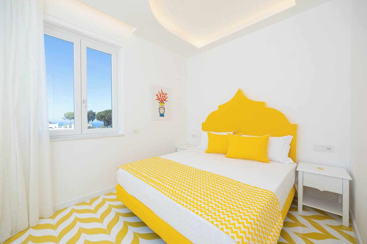 A hotel room with a bed, a window, a nightstand, and a wall-mounted artwork.