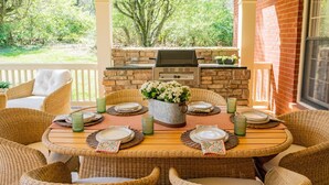 Ein überdachter Patio mit Backsteinmauer, Grill, Korbmöbeln und Blick ins Grüne.