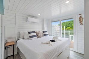 A bedroom with a large bed, white walls, an air conditioning unit, a wooden side table, and a view of a lake and greenery.