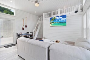 A spacious living area with a white sofa, a dining table with chairs, a staircase, and a large window.