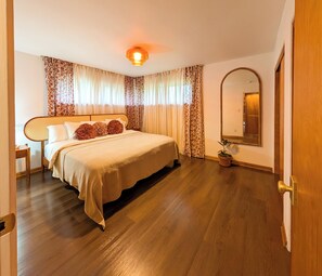 Une chambre à coucher avec un lit, un plancher en bois, des rideaux, un miroir et une porte.