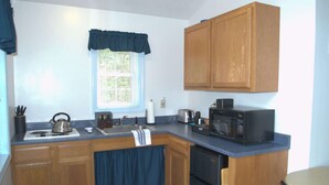 A compact kitchen with wooden cabinets, a sink, a stove, a microwave, and a small dining area. - Little Acorn Landing (Norway)
