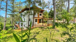 A two-story house with a balcony, surrounded by trees and a grassy yard. - Escápate al Bosque · Villa Completa · Piscina · BBQ · Hasta 12 Huéspedes (Jarabacoa)