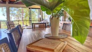 A wooden dining table with a large potted plant, a wooden cutting board, and wicker chairs. - Escápate al Bosque · Villa Completa · Piscina · BBQ · Hasta 12 Huéspedes (Jarabacoa)