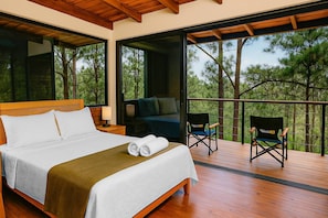 A bedroom with a large bed, a balcony with a view of trees, and a director's chair.
