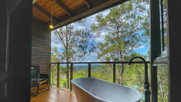 A modern bathroom with a freestanding bathtub, wooden deck, and a view of trees and hills. - Escápate al Bosque · Villa Completa · Piscina · BBQ · Hasta 12 Huéspedes (Jarabacoa)