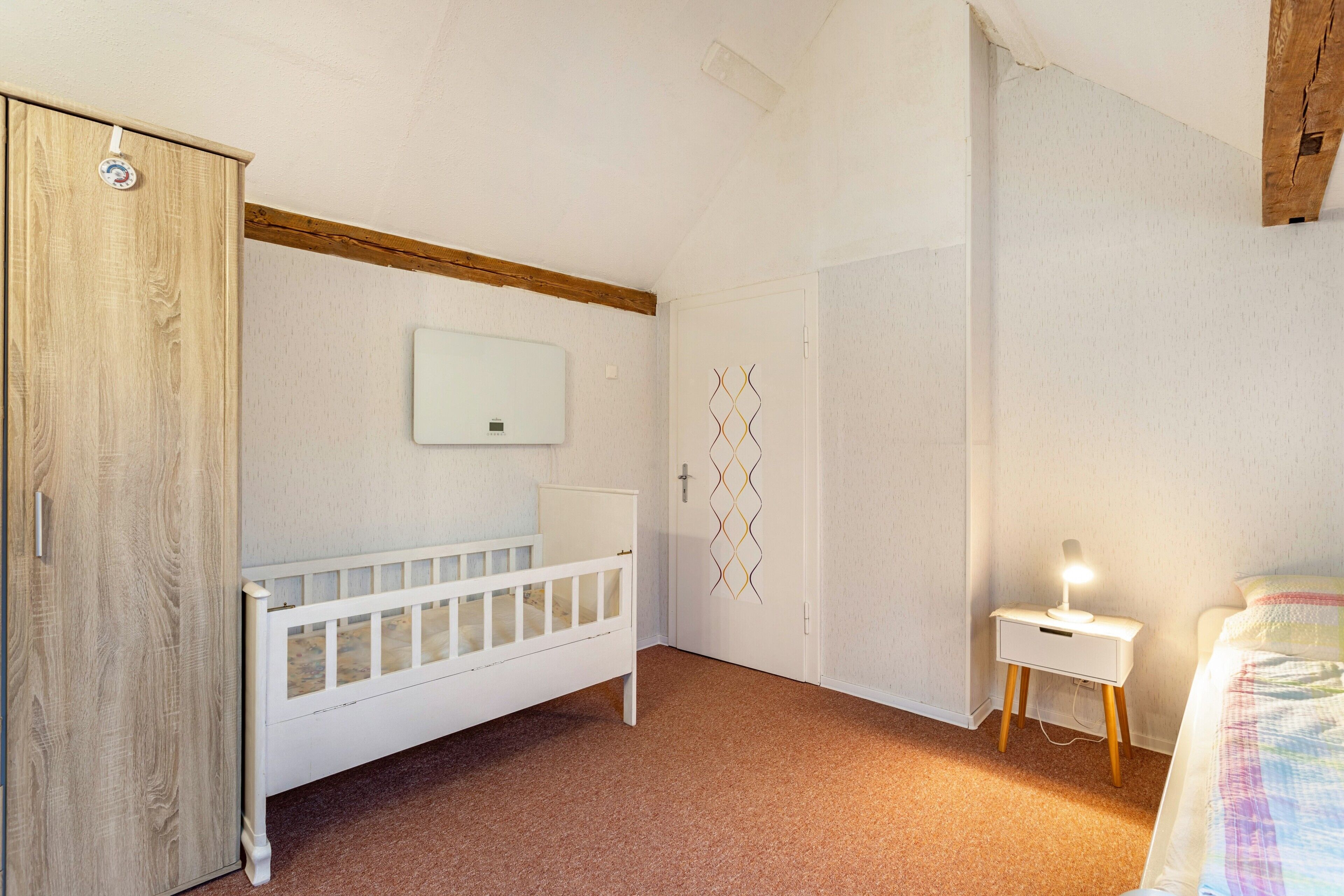 A bedroom with a wooden wardrobe, a white crib, a bedside table with a lamp, and a wall-mounted air conditioner.