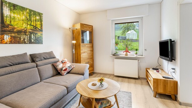 A modern living room with a sofa, a round wooden table, a TV, and a window with a view of greenery. - Apartment "Gerke" with Wi-Fi (Warstein)