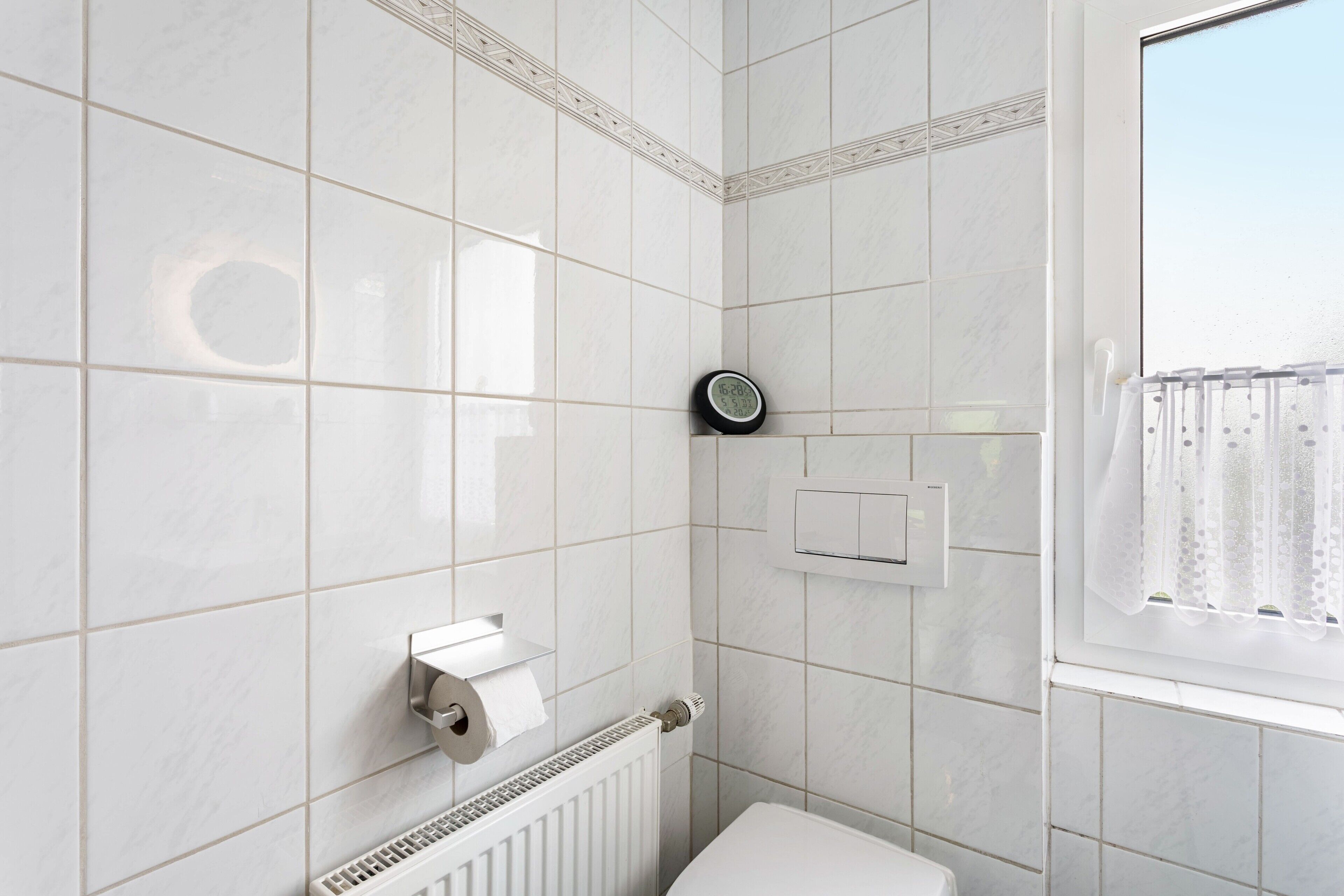 Een badkamer met witte tegels, een toilet, een radiator, een klok en een raam met gordijn.