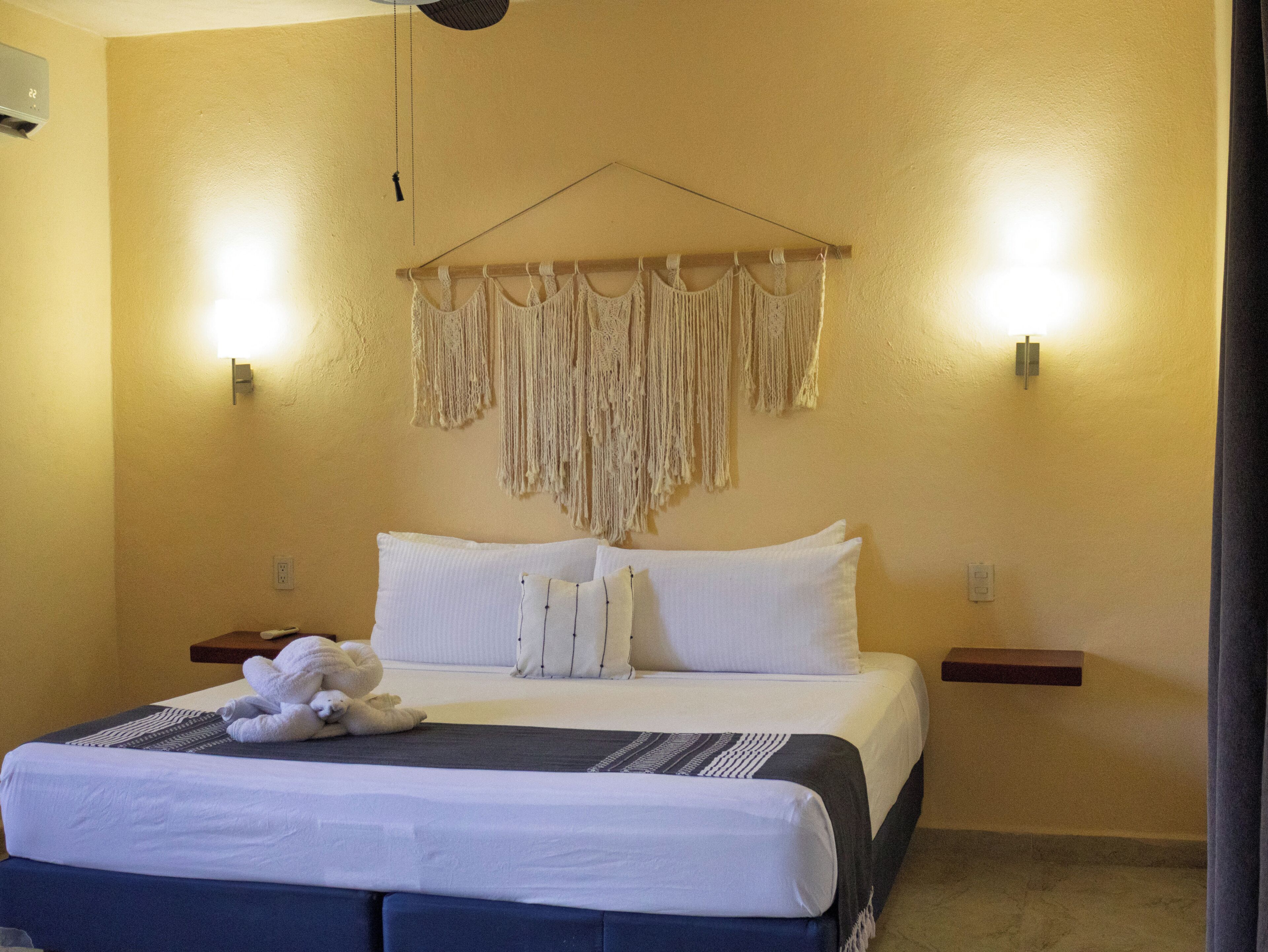 Une chambre d’hôtel avec un lit, des oreillers, une décoration murale en macramé, des lampes de chevet et une petite table de nuit en bois.