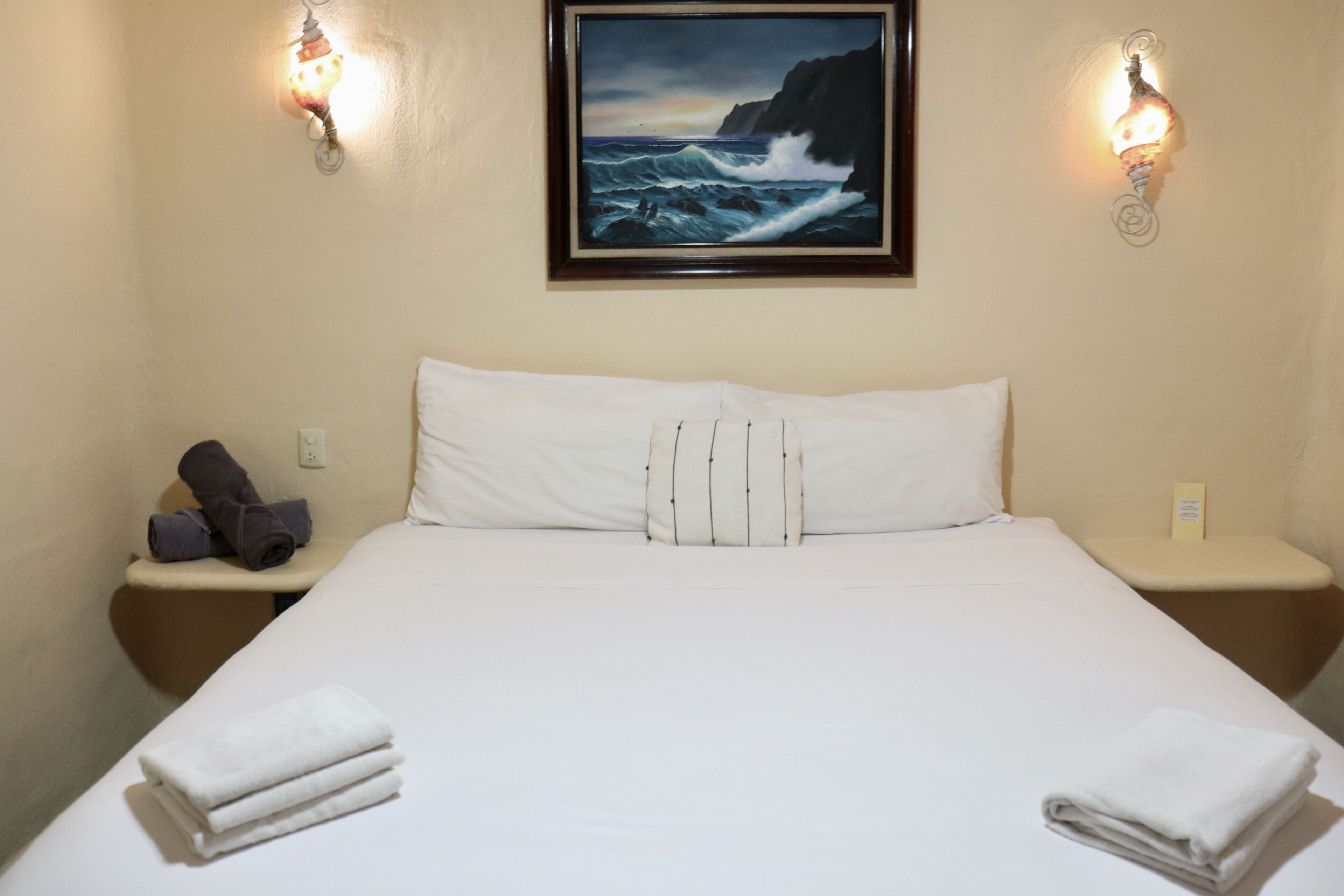 Een hotelkamer met een bed, twee nachtkastjes, een schilderij van de oceaan en wandlampen.