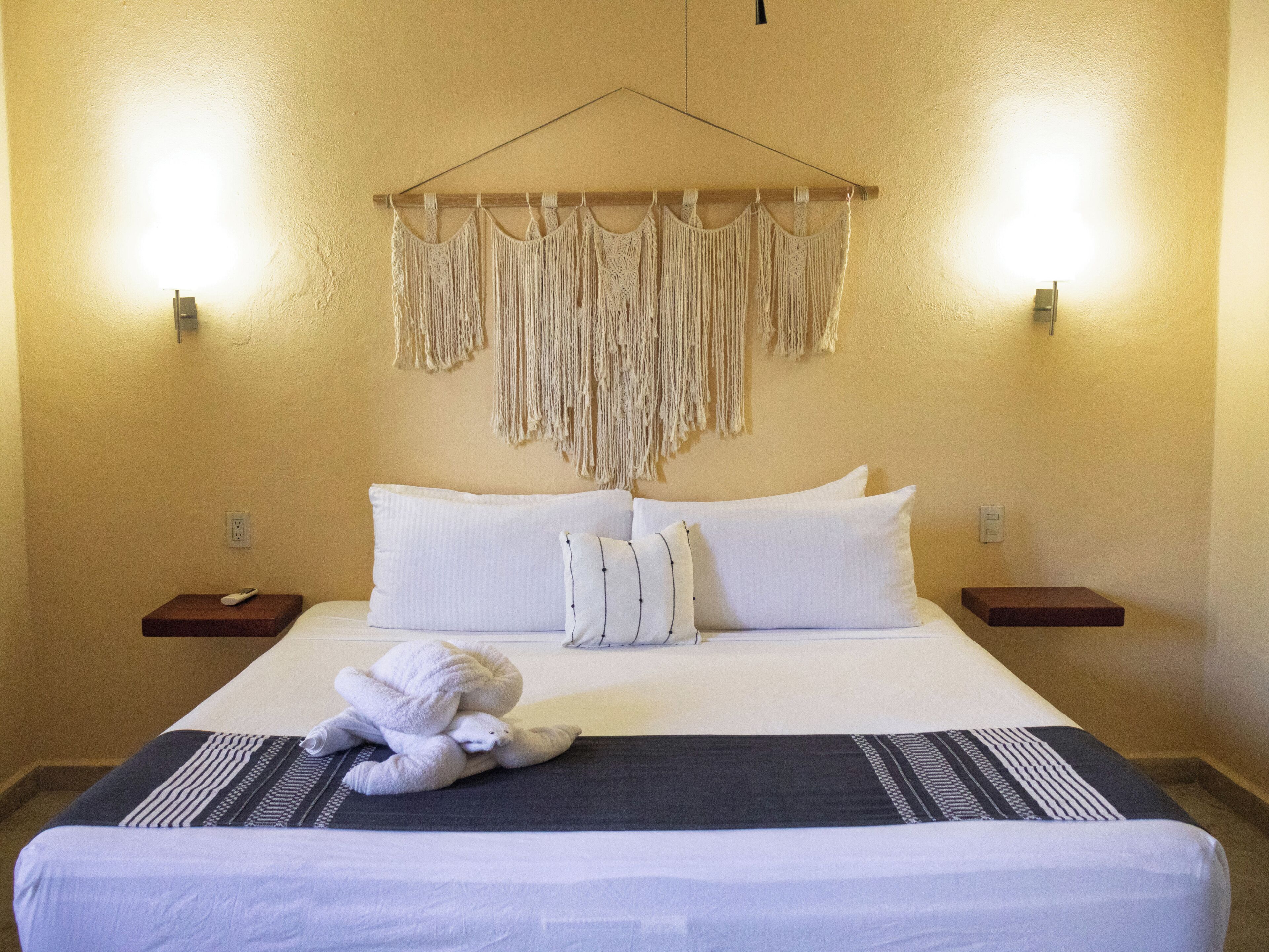 Une chambre d’hôtel avec un lit, des oreillers, une décoration murale en macramé, des tables de chevet et une télécommande.