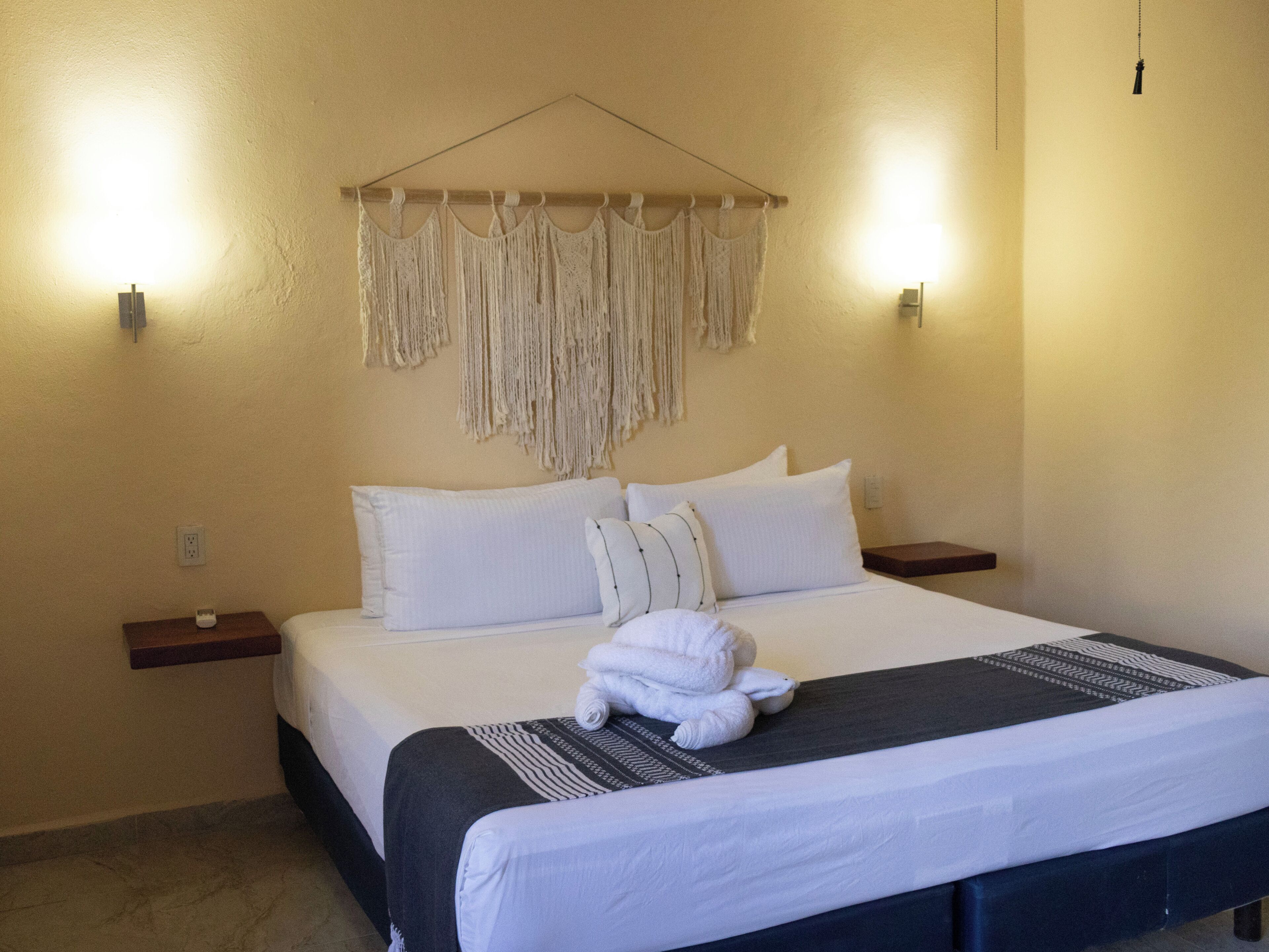 A hotel room with a bed, white pillows, a dark headboard, a macrame wall hanging, bedside tables, and a hanging light fixture.