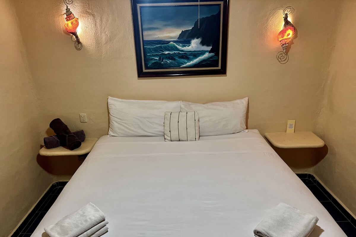 A hotel room with a large bed, two nightstands, a painting of a seascape, and two wall-mounted lamps.