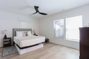 Une chambre à coucher comprenant un lit, des tables de chevet, un ventilateur de plafond, une fenêtre avec des stores et une vue sur un bâtiment à l’extérieur.