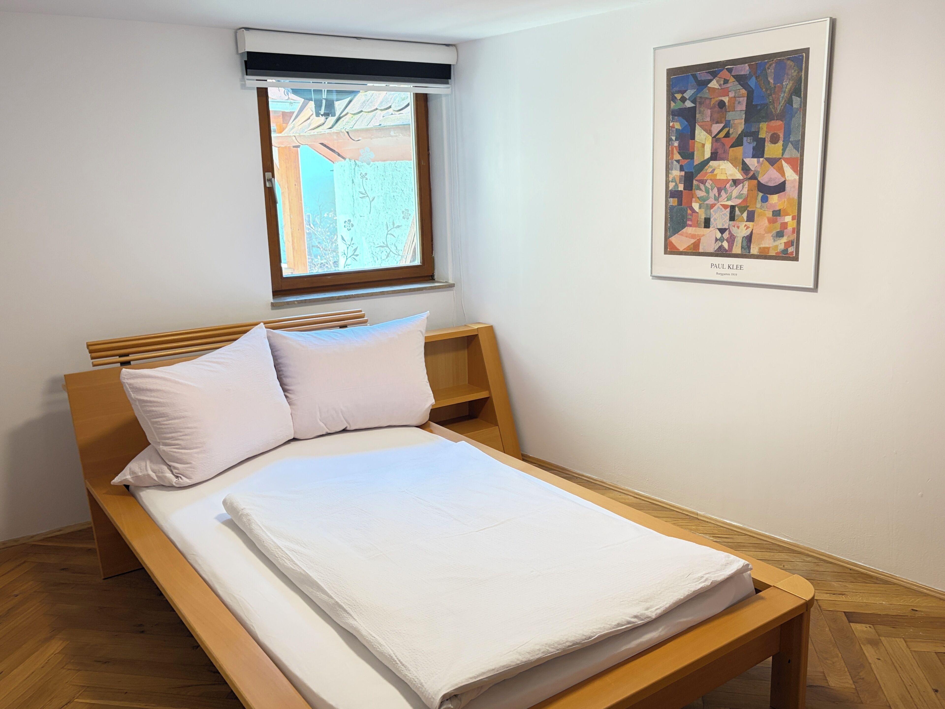 A single bed with a wooden frame and white bedding, a window with a view of greenery, and a framed abstract painting on the wall.