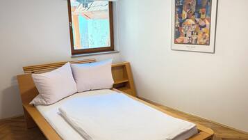 A single bed with a wooden frame and white bedding, a window with a view of greenery, and a framed abstract painting on the wall.