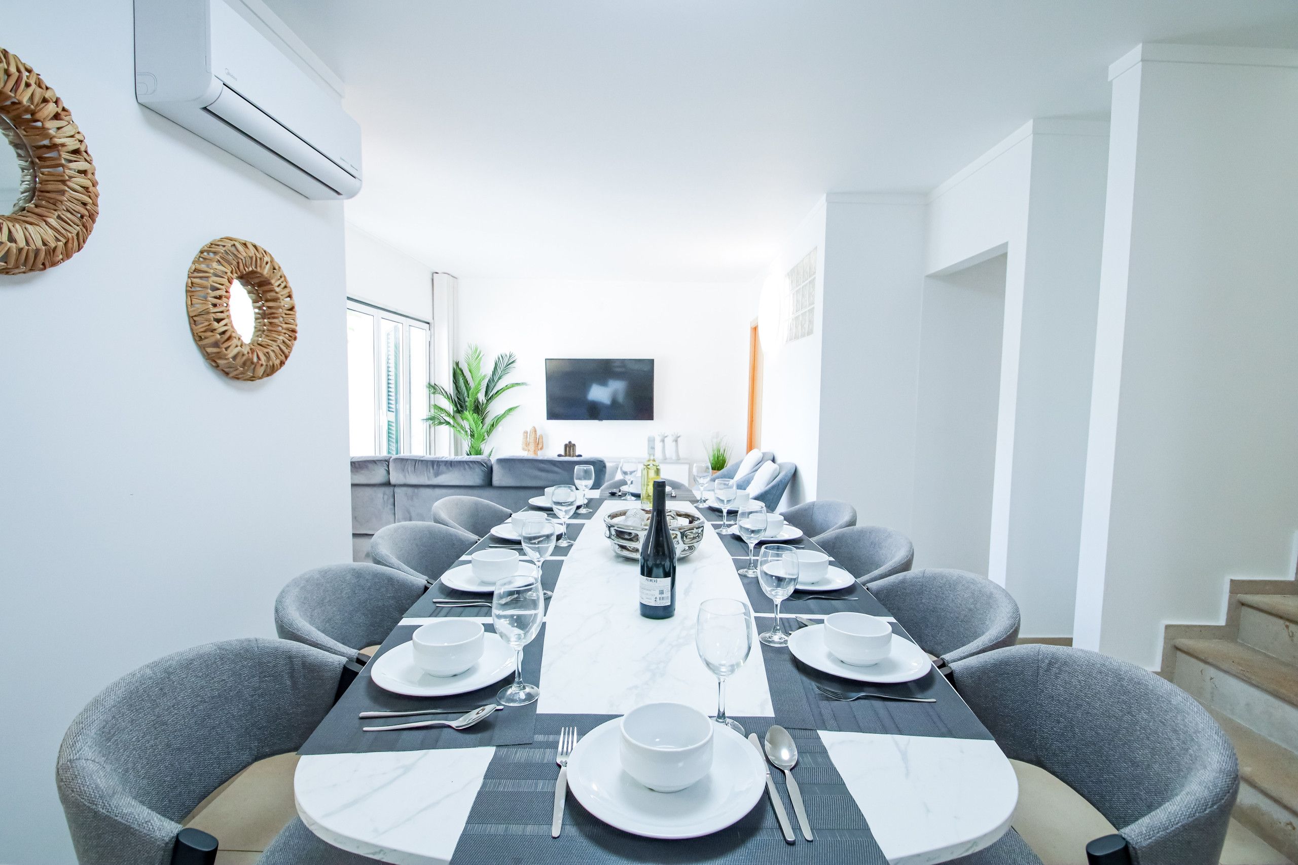 A modern dining room with a long table set for a meal, grey upholstered chairs, and a flat-screen TV mounted on the wall.