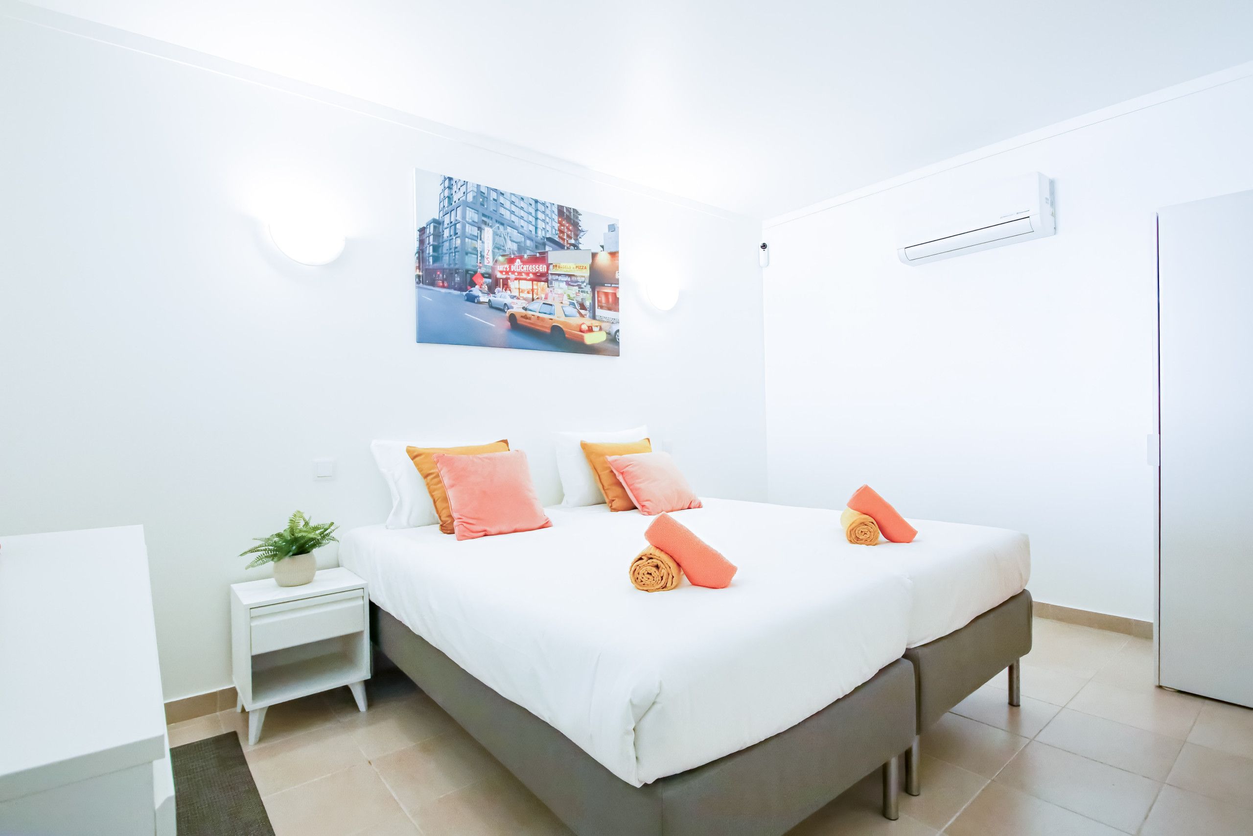 A modern hotel room with a bed, a nightstand, a small plant, and a wall-mounted air conditioning unit.