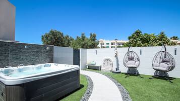 A backyard with a hot tub, two egg chairs, a stone bench, and a stone wall.
