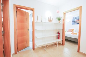 A hotel room with a wooden door, a white shelving unit with decorative items, a bed with pillows, and a fire extinguisher.