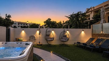 A hot tub, two hanging chairs, and lounge chairs in a backyard with a sunset view.