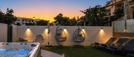 Ein Whirlpool, zwei Hängesessel und Liegestühle in einem Garten mit Sonnenuntergangsblick.