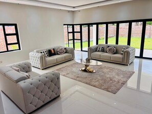 A spacious living room with two sofas, a coffee table, and large windows offering a view of a brick wall and greenery. - Spacious 5-bedroom house in charming Harare with AC (Harare)