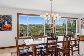 A dining area with a large table and chairs, a chandelier, and a view of a balcony with plants and a railing.