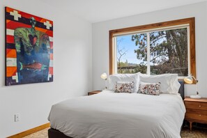 Un dormitorio con una cama grande, dos mesitas de noche, un ventanal con vistas a árboles y un cuadro abstracto y colorido en la pared.