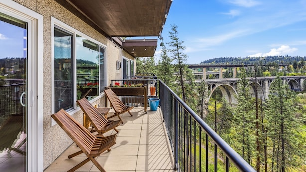 A balcony with wooden chairs, a blue trash bin, and a view of a bridge and forest.