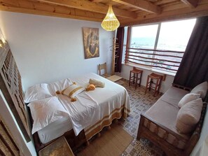 Una habitación con cama, sofá, piso de madera, ventana con vistas y lámpara colgante.