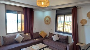 Ein Wohnzimmer mit einem Ecksofa, einem Couchtisch, einem Fernseher und einem großen Fenster mit Vorhängen.