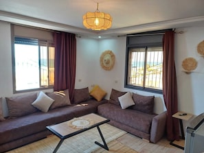 Una sala de estar con un sofá modular, una mesa de centro, un televisor y un amplio ventanal con cortinas.
