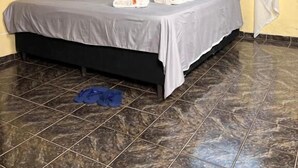 Una cama individual con colcha gris, dosel blanco, aire acondicionado de pared, tapiz de macramé y suelo de baldosas.