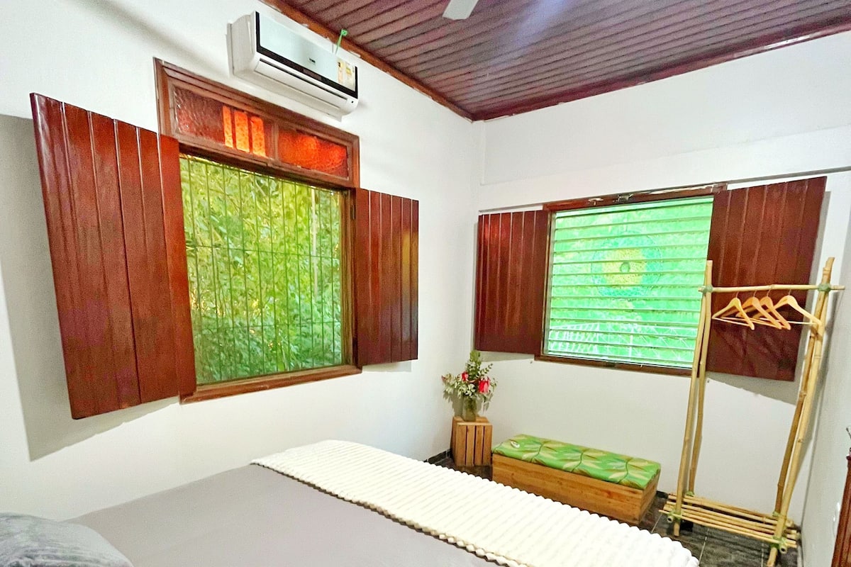 Un dormitorio con cama, ventilador de techo, equipo de aire acondicionado, ventana con contraventanas de madera, pequeña mesa con un florero y un tendedero de madera.
