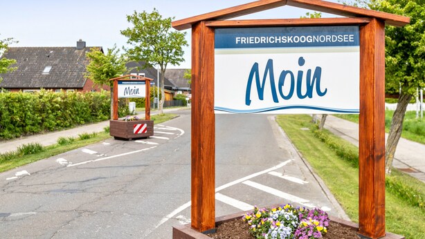 Segno per Friedrichskoog Noordzee con la scritta 'Moin', una struttura in legno e aiuola fiorita.