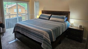 Quarto com cama, ventilador de teto, ar condicionado, mesa de cabeceira e vista para um deck de madeira.