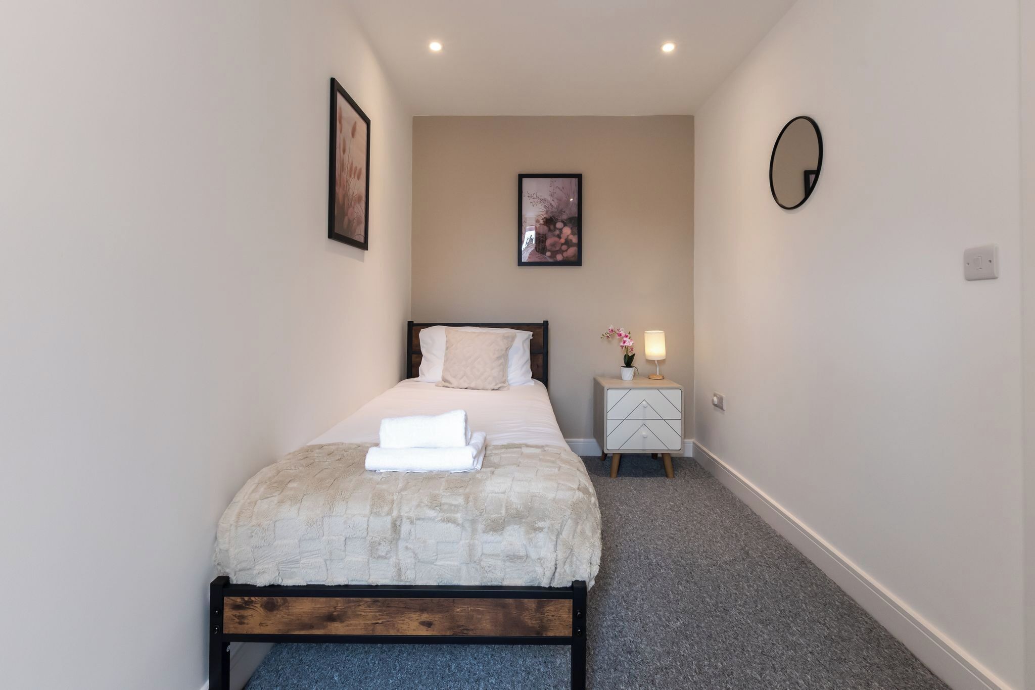 Een slaapkamer met een bed, nachtkastje, lamp, een lijst met afbeelding en een spiegel.